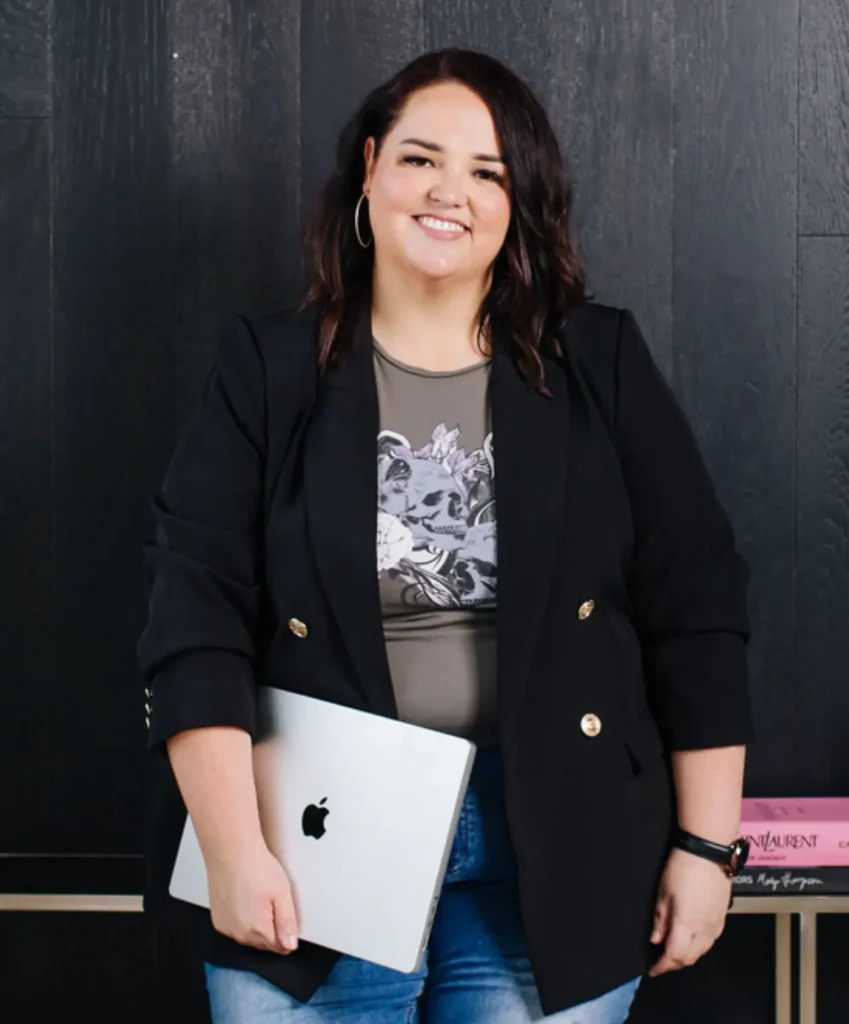 Jemimah Ashleigh wearing a dark top, black blazer and jeans. She is leaning against a gold table in front of a black wall while holding her closed laptop against herself. She has wavy brown hair and is smiling.