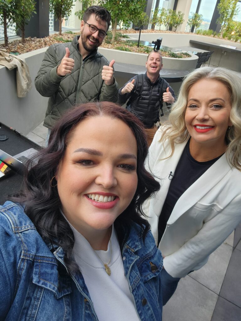 A selfie of Jemimah and Danica with the team from Do Good Studios posing with "thumbs up" behind them. Jemimah has dark wavy hair and wears a denim jacket, Danica has blonde wavy hair and wears a white blazer. The two men in the background have dark coloured winter coats.