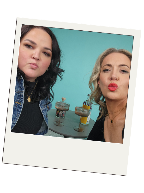 Jemimah and Danica blowing kisses at the camera. Jemimah has wavy brown shoulder length hair and Danica has wavy long blonde hair. Behind them is a table with cocktails.